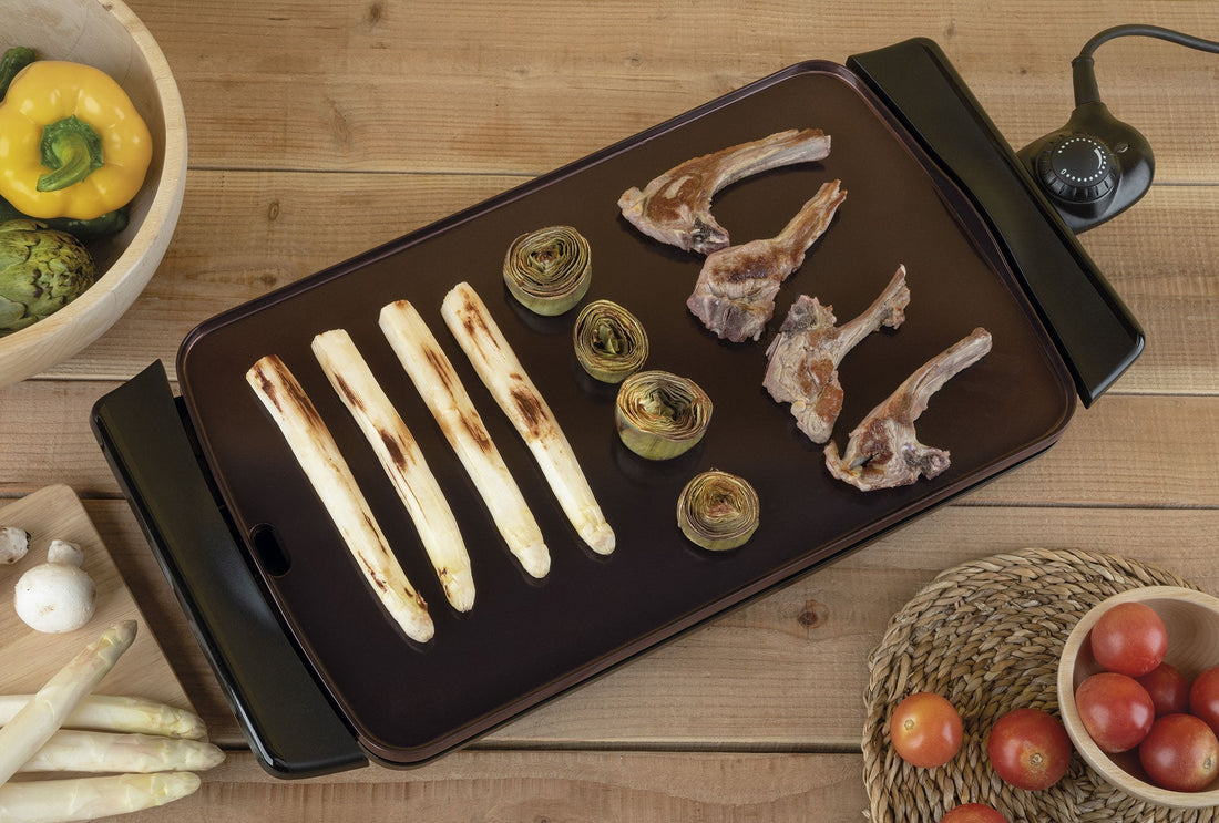 Parrilla eléctrica con espárragos, alcachofas y costillas cocinándose, alrededor verduras frescas.