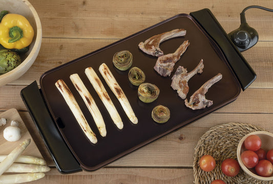 Parrilla eléctrica con espárragos, alcachofas y costillas cocinándose, alrededor verduras frescas.